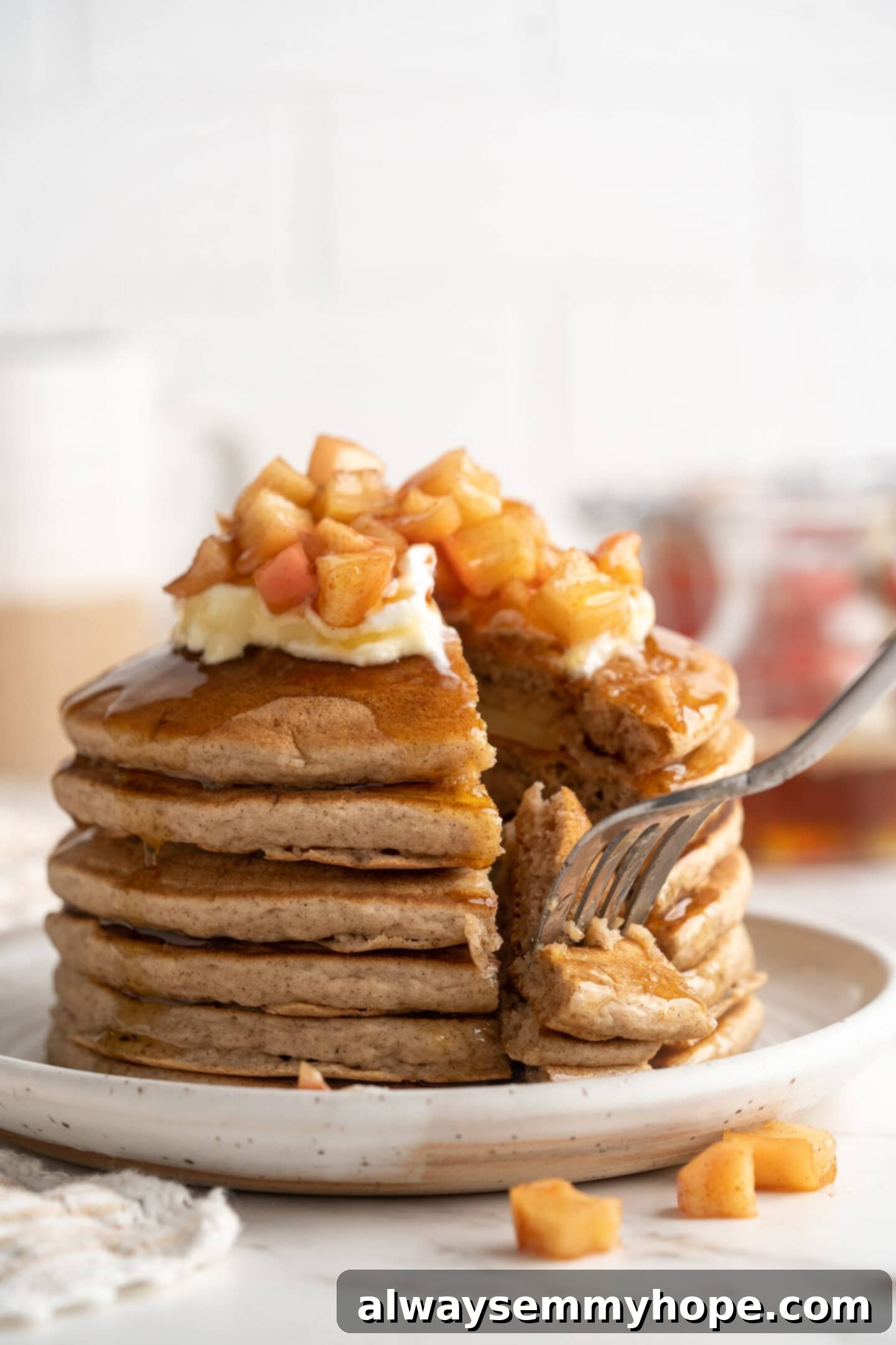 A fork is delicately piercing a bite-sized piece of a fluffy vegan apple pie pancake, showing the tender interior and the embedded apple slices, ready to be enjoyed. The shot emphasizes the inviting texture and deliciousness.