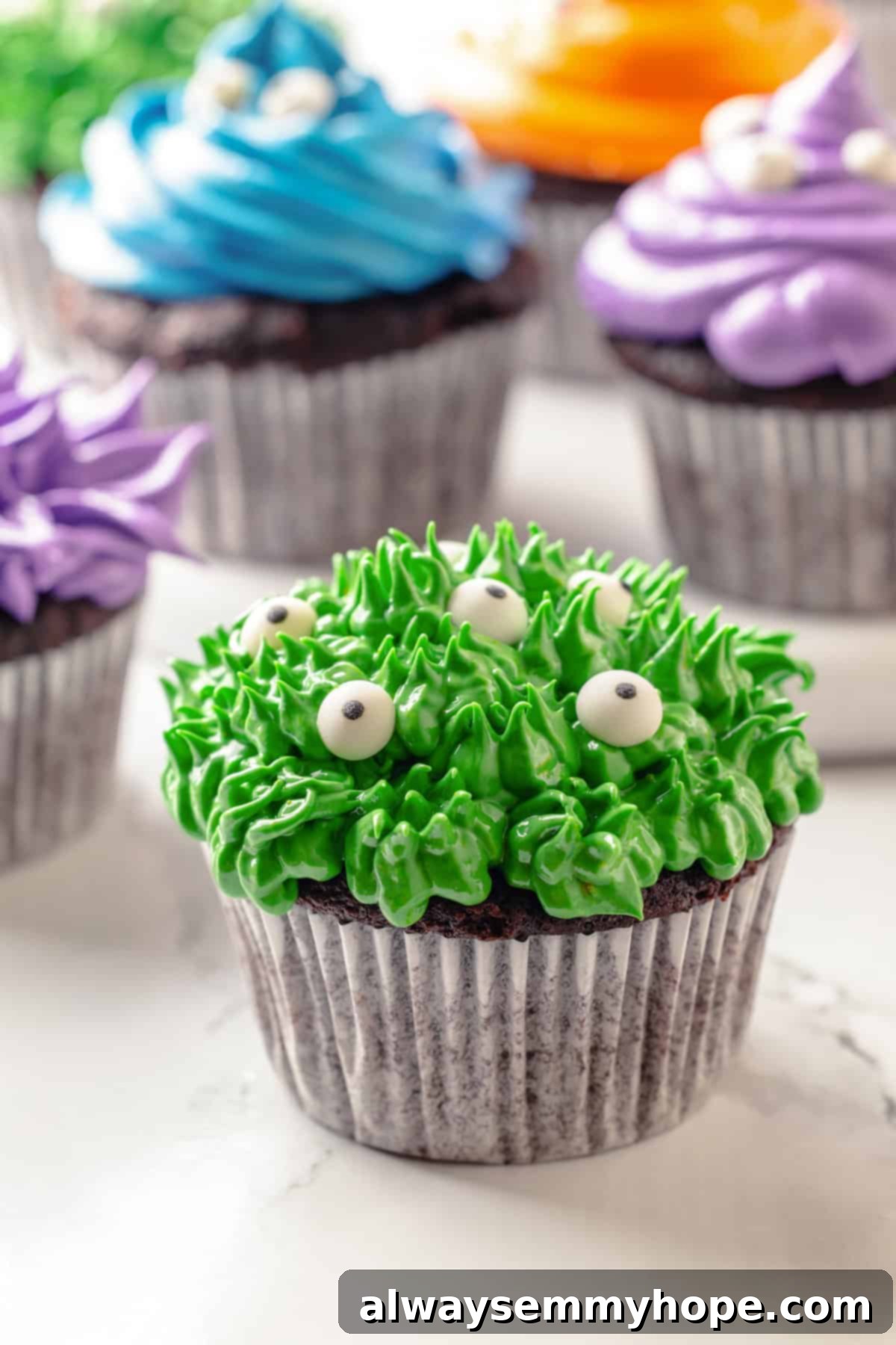These moist chocolate vegan Halloween cupcakes are topped with cute frosting monsters in a variety of colours, making them a festive and delicious plant-based dessert for any spooky celebration. Vibrant green furry monster Halloween cupcake, with other colorful monster cupcakes blurred in the background, showcasing the playful design and delicious appeal of these vegan treats.