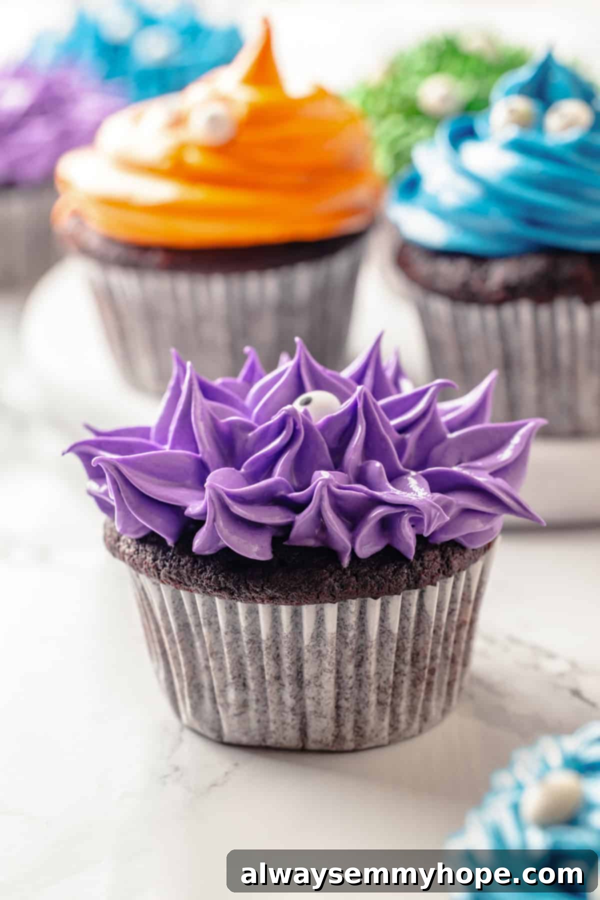 These moist chocolate Halloween cupcakes are topped with cute frosting monsters in a variety of colours. They taste as good as they look! Close-up of a purple furry monster Halloween cupcake with candy eyes, blurred background of other monster cupcakes, emphasizing its cute and festive appearance.