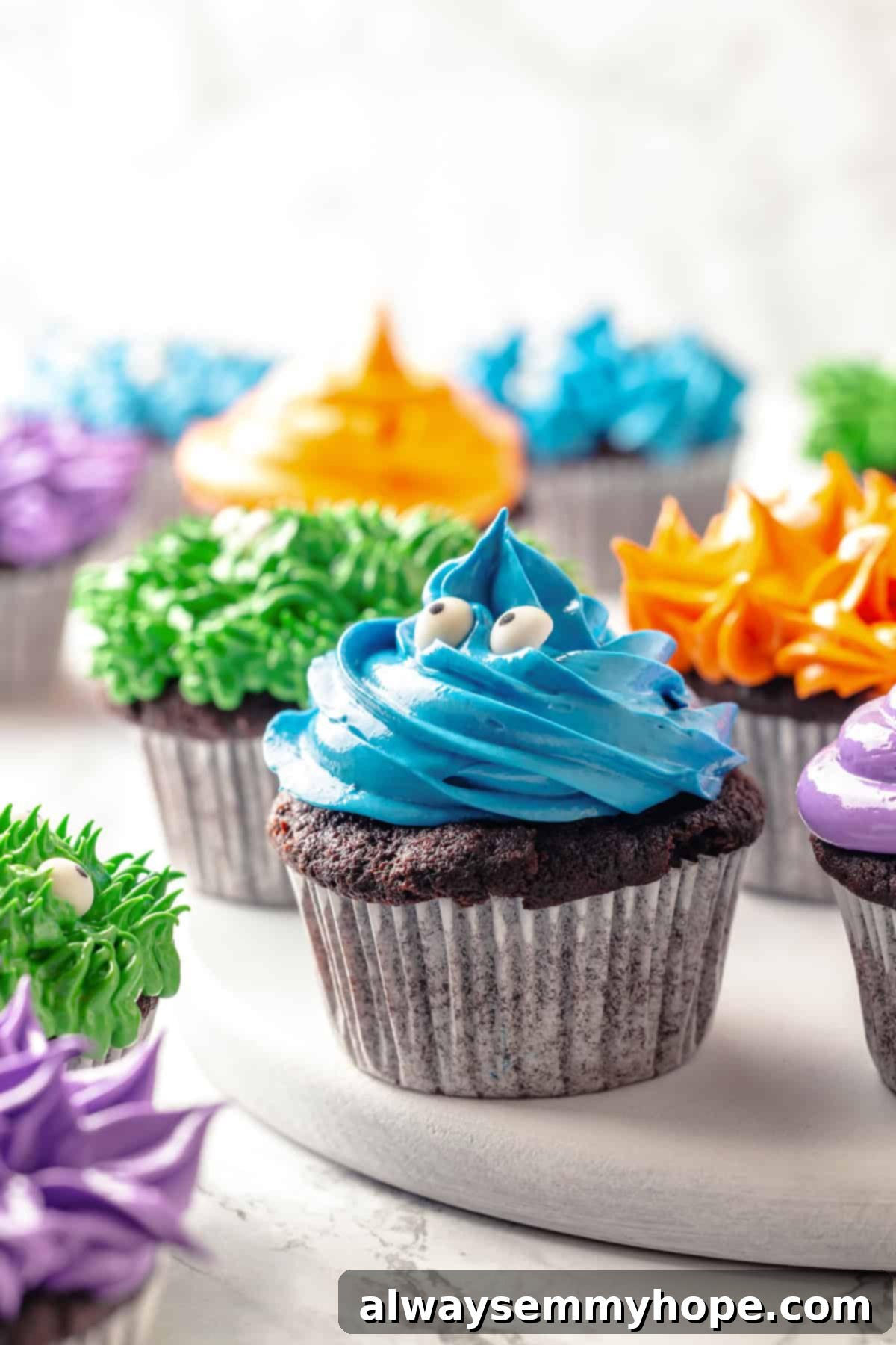 These moist chocolate Halloween cupcakes are topped with cute frosting monsters in a variety of colours. They taste as good as they look! Close-up of a blue furry monster Halloween cupcake with candy eyes, with other colorful monster cupcakes in soft focus in the background, highlighting the fun decoration.