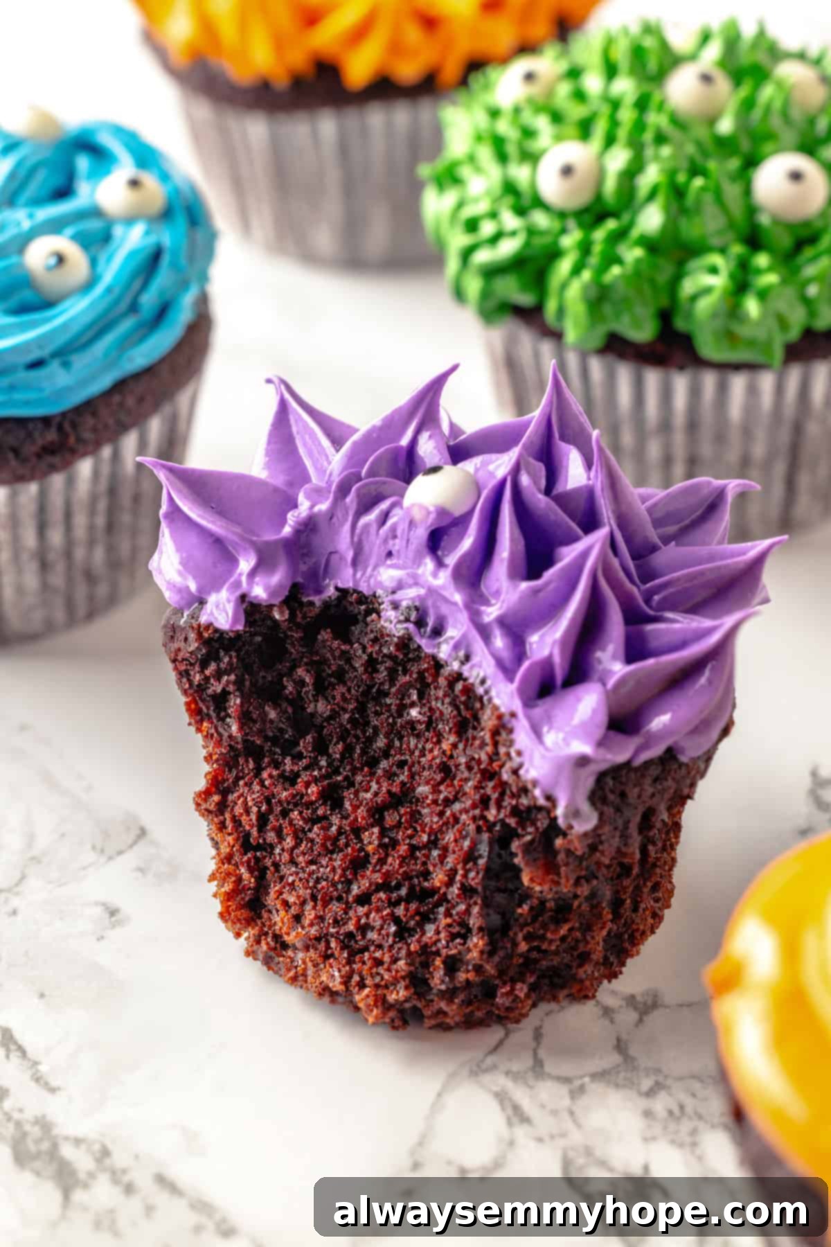 These moist chocolate Halloween cupcakes are topped with cute frosting monsters in a variety of colours. They taste as good as they look! A purple monster Halloween cupcake with a bite taken out, revealing the fluffy chocolate cake texture beneath the furry frosting and candy eyes.