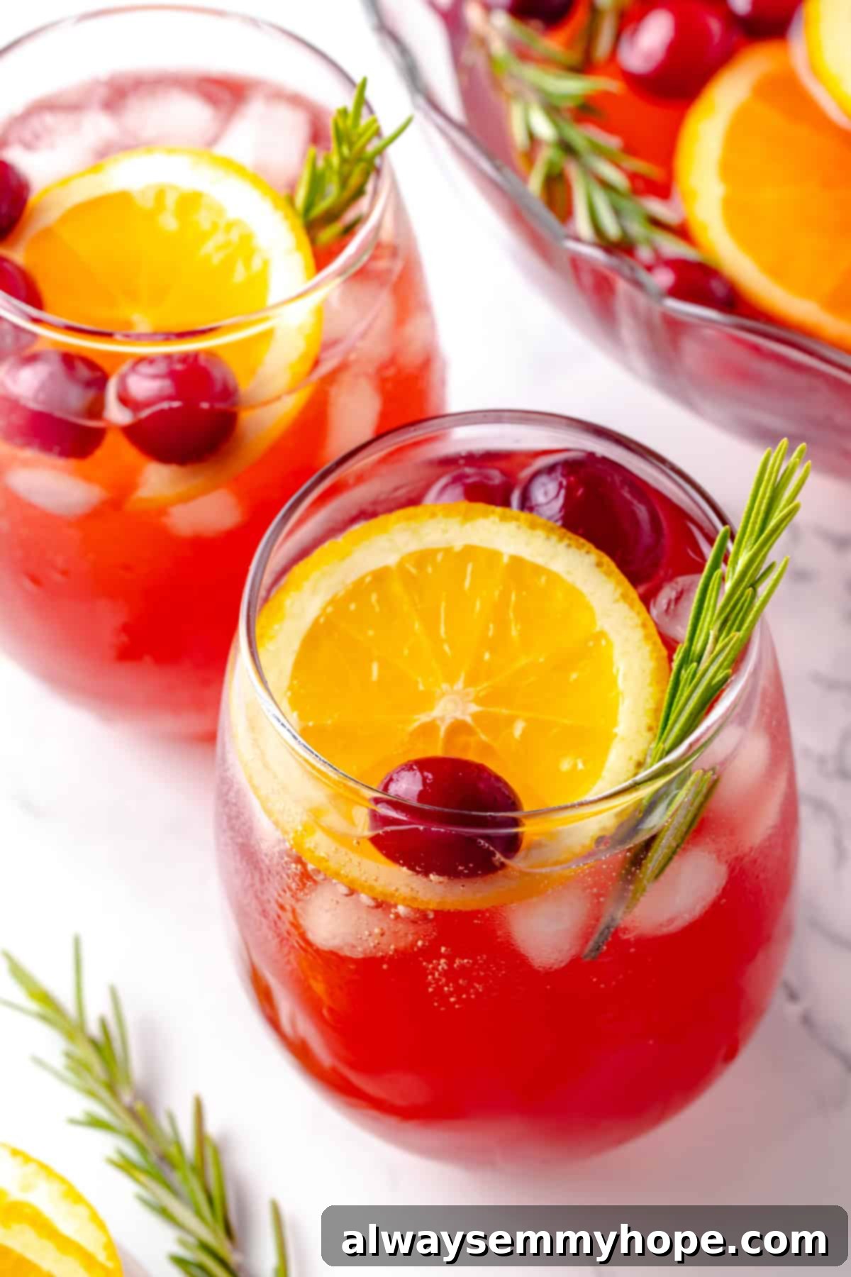 Two elegant glasses of Christmas punch, garnished with vibrant orange slices, fresh rosemary, and a sprinkle of cranberries, set against a festive background.