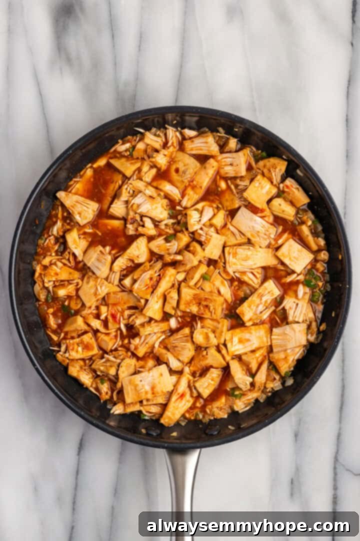 Overhead view of sliced jackfruit simmering in a rich, spiced sauce with vegetable broth, lime juice, tomato paste, and soy sauce in a skillet, covered to infuse flavors.
