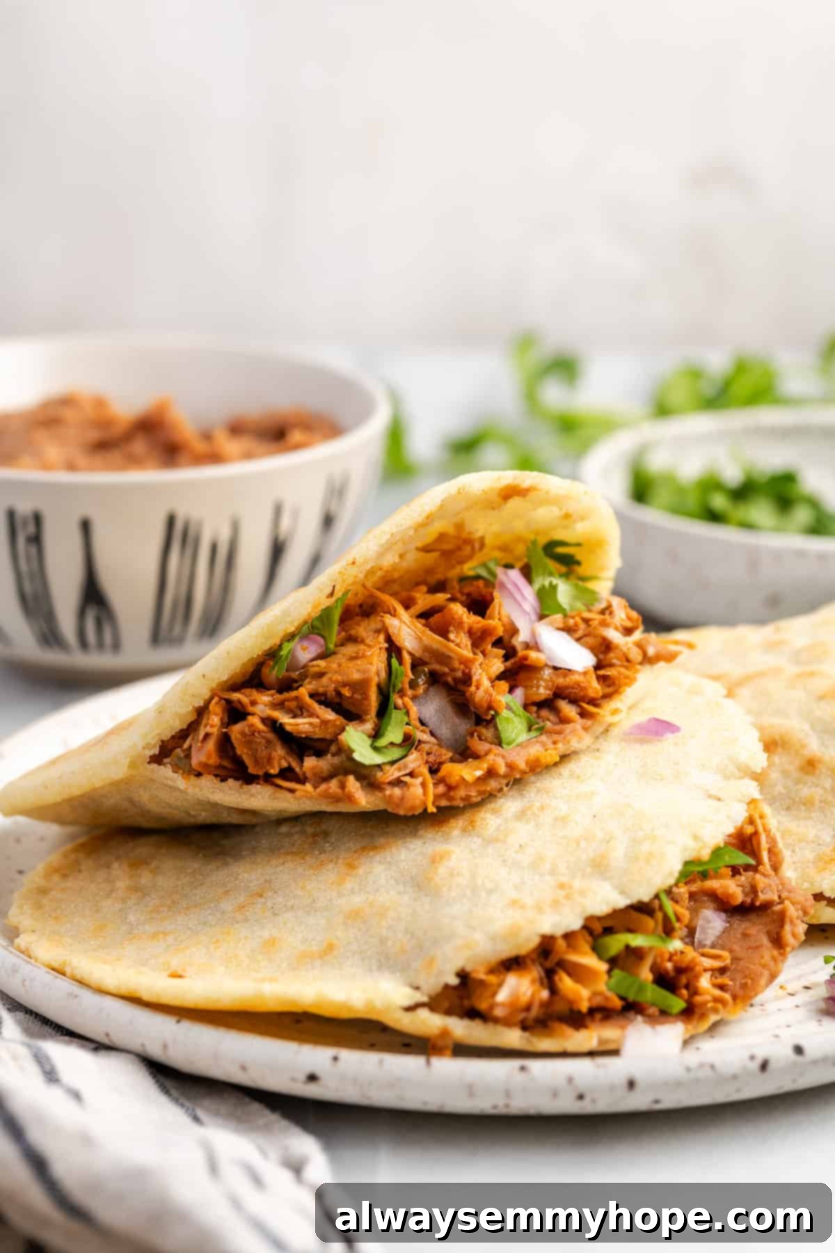 A single homemade vegan jackfruit gordita, cut open and generously stuffed with shredded jackfruit, fresh cilantro, and a drizzle of sauce, resting on a white plate with lime wedges.