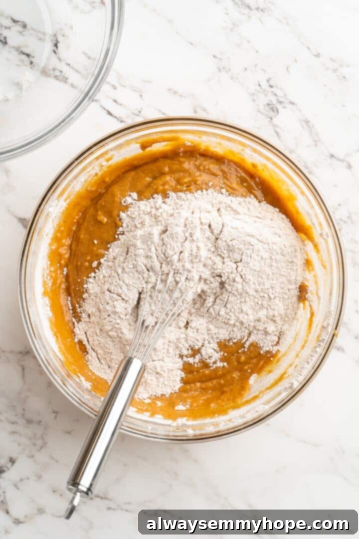 Vegan pumpkin bread with cream cheese frosting is moist, full of cozy spices and the frosting takes it over the top. So easy to make too! Dry ingredients for pumpkin bread are being poured into a bowl containing the mixed wet ingredients, showing the two mixtures about to be combined.