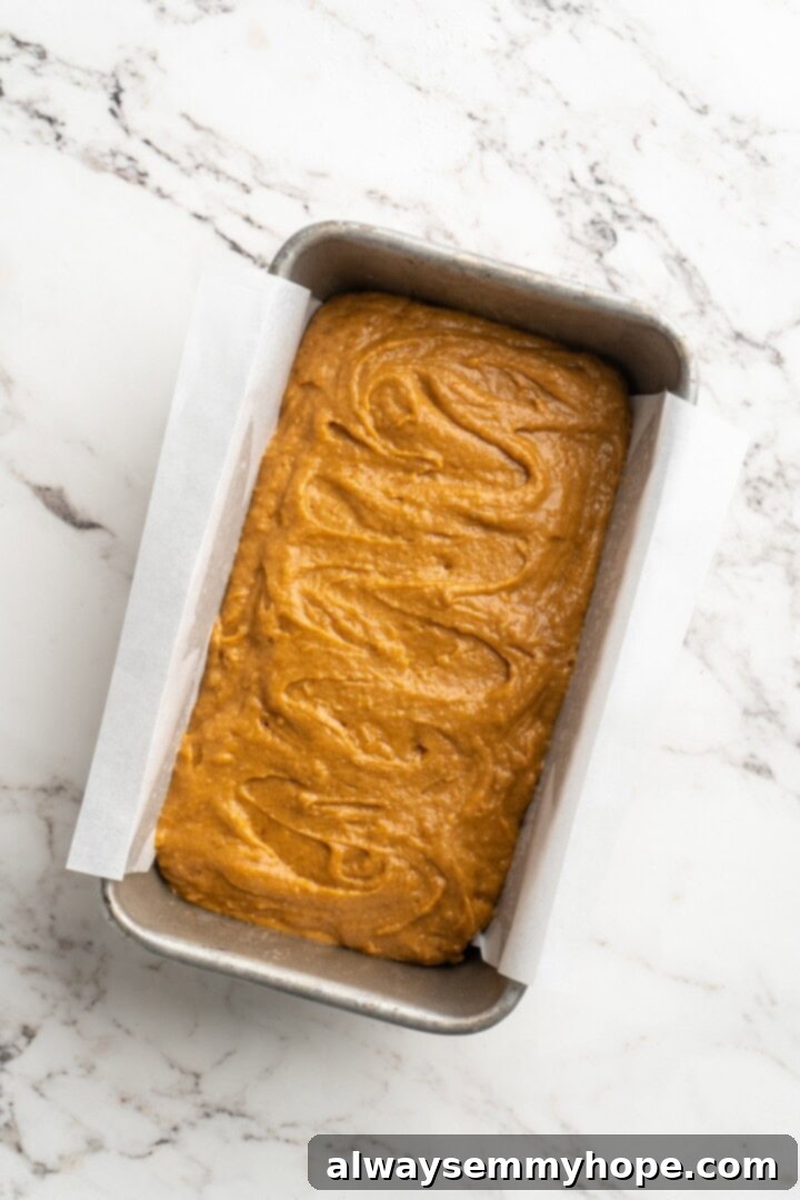 Vegan pumpkin bread with cream cheese frosting is moist, full of cozy spices and the frosting takes it over the over the top. So easy to make too! Overhead view of thick pumpkin bread batter being spread evenly into a parchment-lined loaf pan, ready for baking.