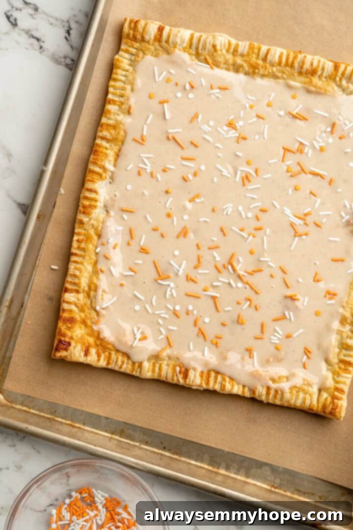 A cozy pumpkin-spiced filling is tucked into flaky vegan puff pastry to make these easy and delicious homemade pumpkin pop tarts! Giant baked pumpkin pop tart on a baking sheet, frosted and ready to be cut into individual servings.
