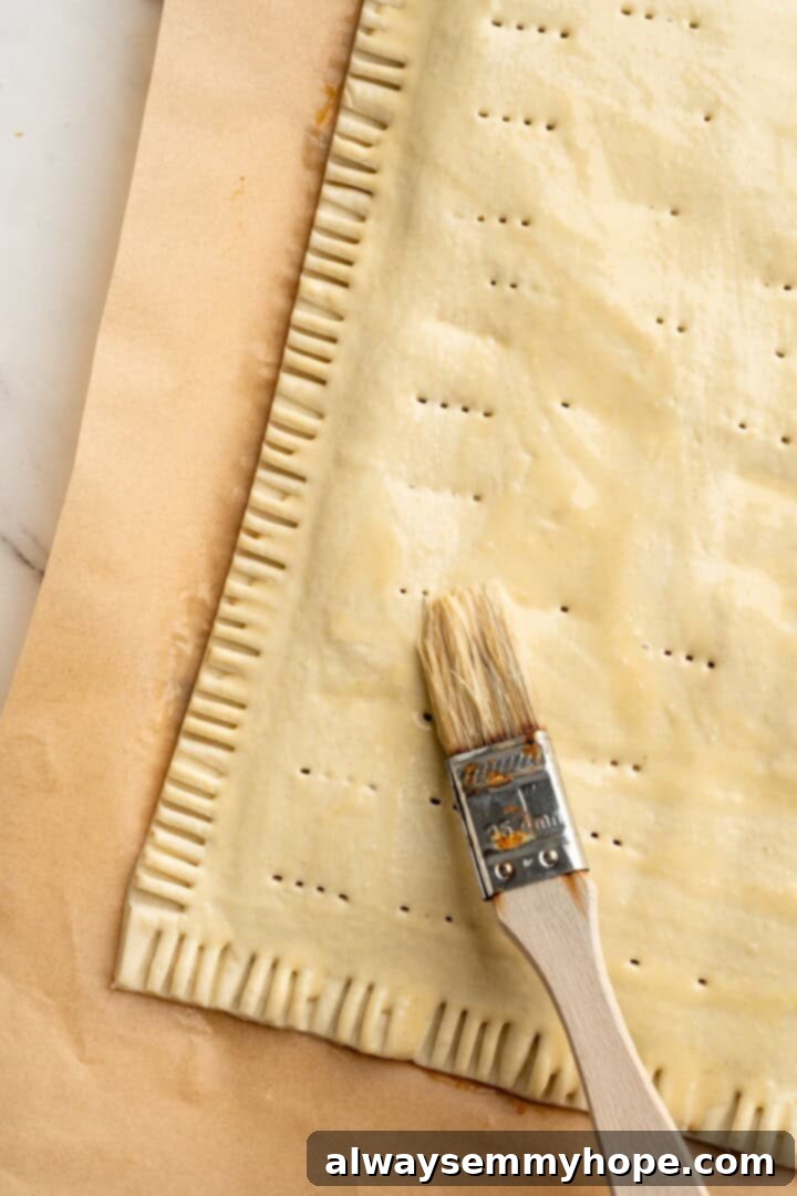 A cozy pumpkin-spiced filling is tucked into flaky vegan puff pastry to make these easy and delicious homemade pumpkin pop tarts! Brushing vegan egg wash over the top of the assembled pumpkin pop tart before baking.