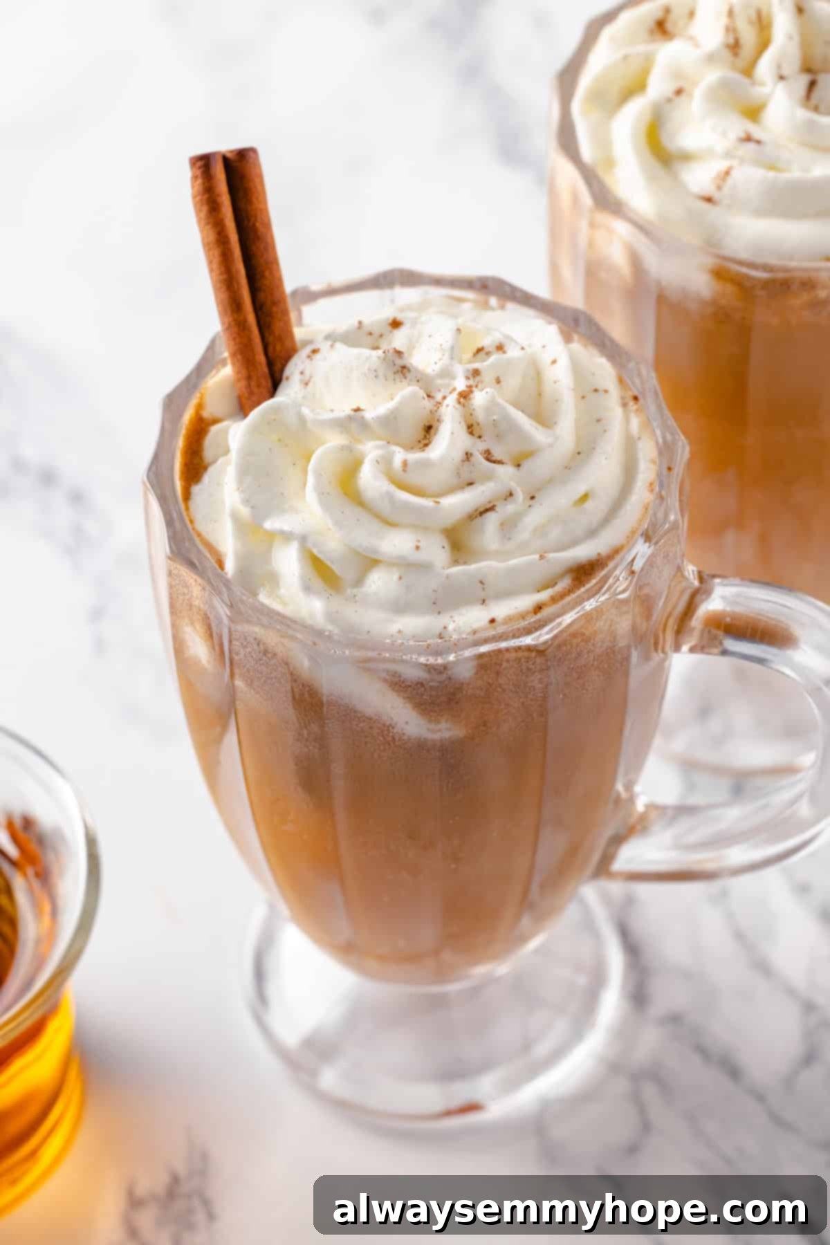 A stunning shot of a single hot buttered rum, complete with whipped cream and cinnamon stick. A single elegant glass mug of hot buttered rum, topped with a swirl of vegan whipped cream and a cinnamon stick, captured against a festive blurred background.