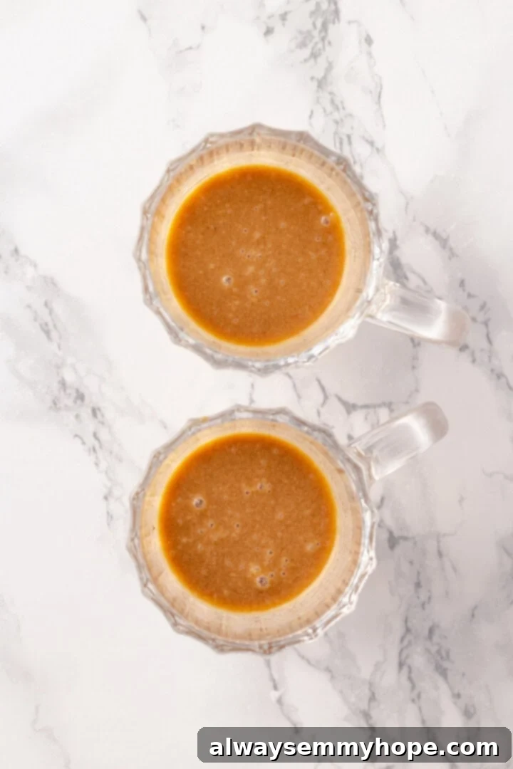 Preparing hot buttered rum by adding the butter base and rum to a mug. Overhead view of two glass mugs with scoops of butter base and rum poured over them, ready for hot water.