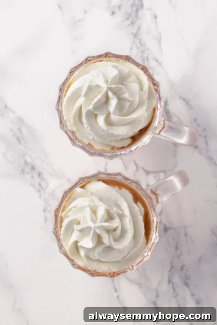 The final touch: garnishing the hot buttered rum with whipped cream and spices for a festive presentation. Overhead view of a finished hot buttered rum cocktail, generously topped with vegan whipped cream and a sprinkle of cinnamon, plus a cinnamon stick garnish.
