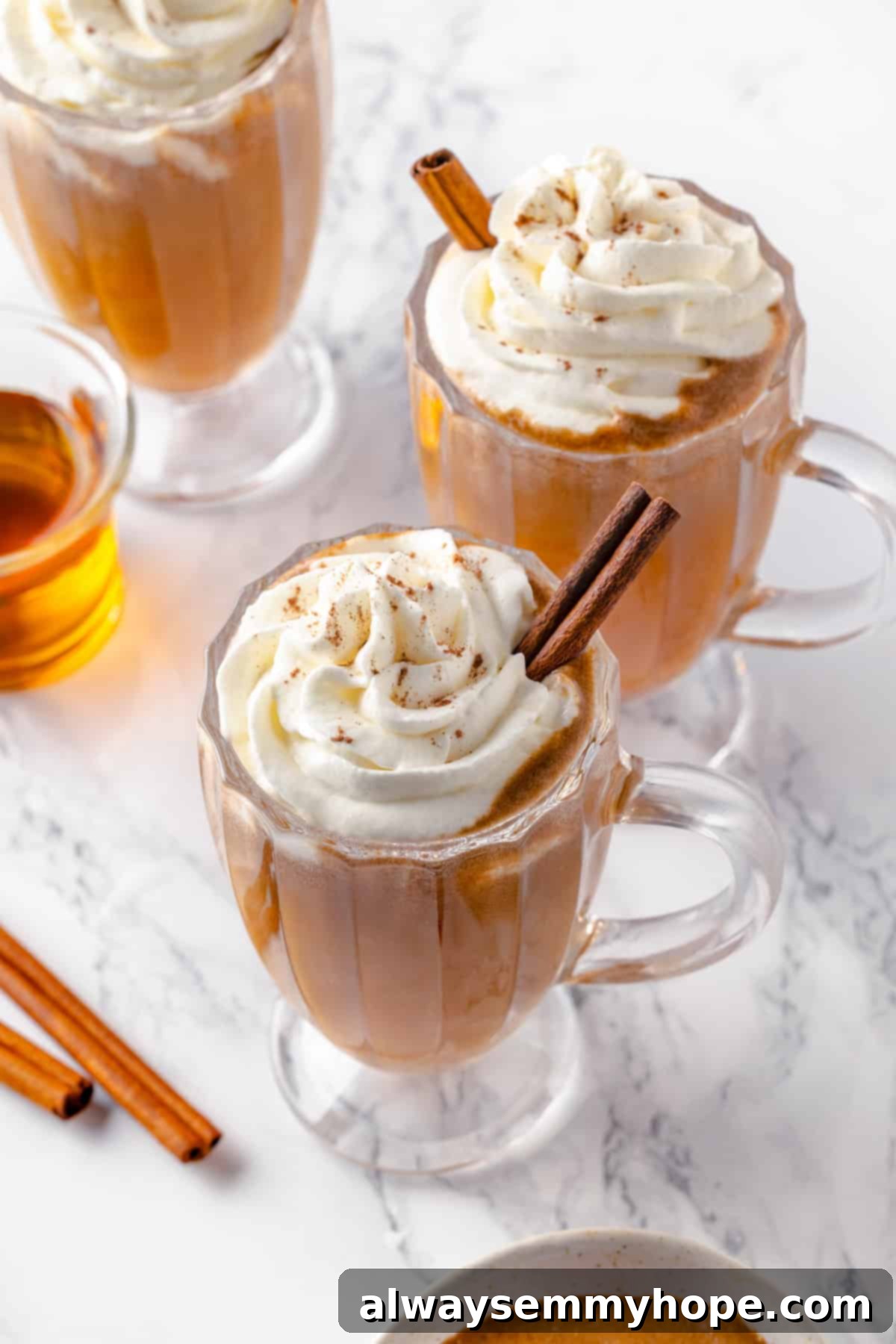 Three mugs of delicious vegan hot buttered rum, beautifully garnished and ready to enjoy. A trio of perfectly prepared glass mugs filled with vegan hot buttered rum, garnished with whipped cream and cinnamon sticks, presented on a rustic surface.