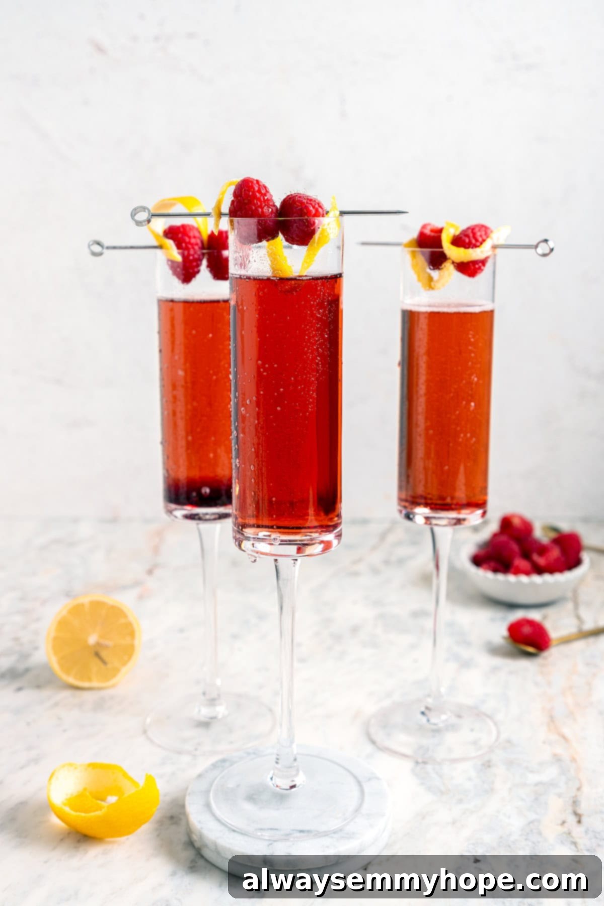 Three perfectly prepared Kir Royale cocktails, each in a champagne flute, garnished with fresh raspberries and a lemon twist, ready to be served.