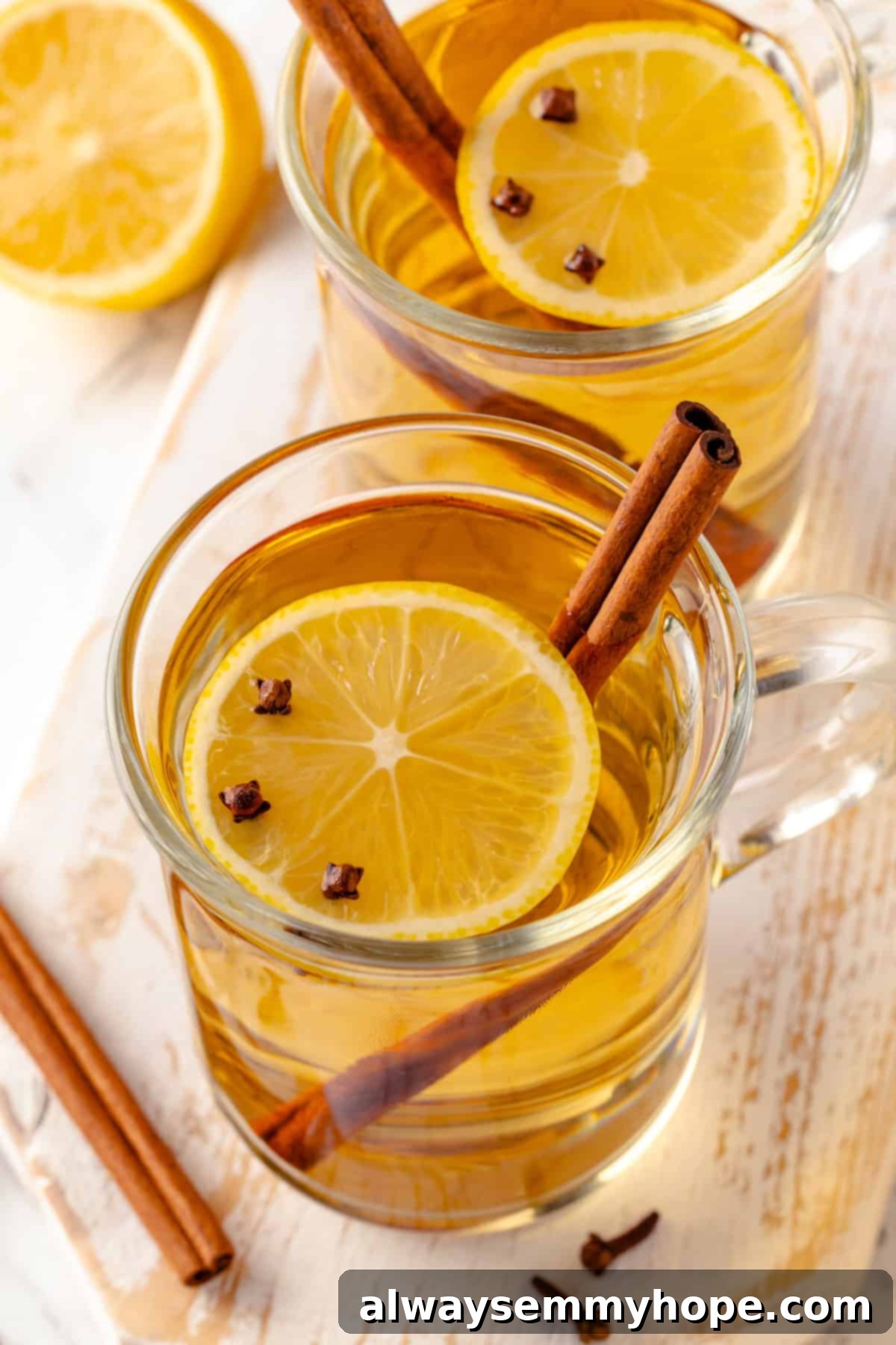 This hot toddy recipe will cure whatever ails you! It's a classic cocktail made with whiskey, lemon juice, and maple syrup to make it vegan. Two mugs of vegan hot toddy, garnished with lemon, cloves, and cinnamon stick, on a wooden surface