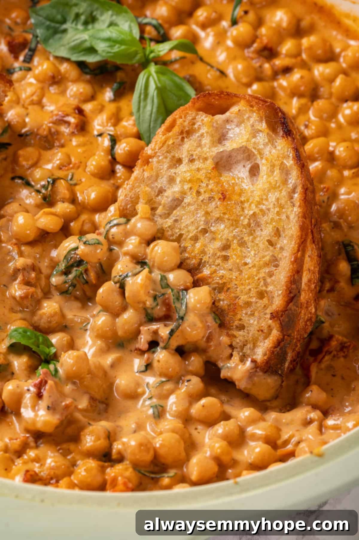 Close-up of crusty bread being dipped into a bowl of creamy Marry Me Chickpeas, showcasing the rich sauce and vibrant ingredients like sun-dried tomatoes and fresh basil.