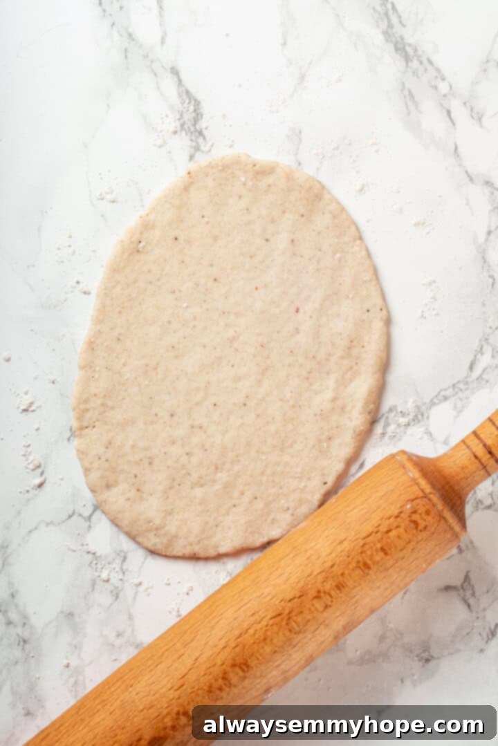 This easy gluten-free naan recipe has the chewy texture and slightly charred bits that make naan so irresistible, but without the wheat! Hands rolling out gluten-free naan dough with a rolling pin on a lightly floured surface, forming an oval shape.