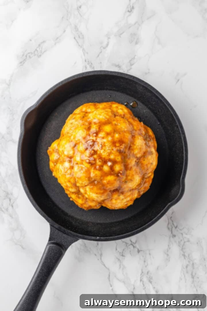 A whole head of cauliflower placed in a cast iron skillet, being brushed generously with the savory butter mixture.