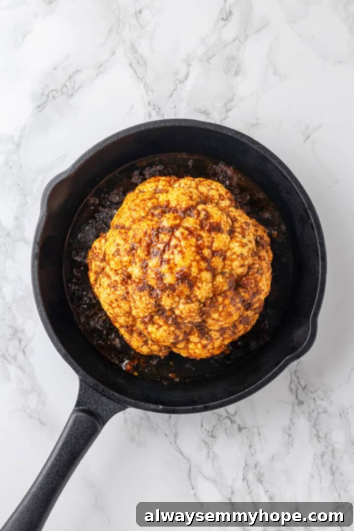 An overhead shot of a whole roasted cauliflower in a cast iron skillet, golden brown and partially cooked, before the final butter and cheese application.