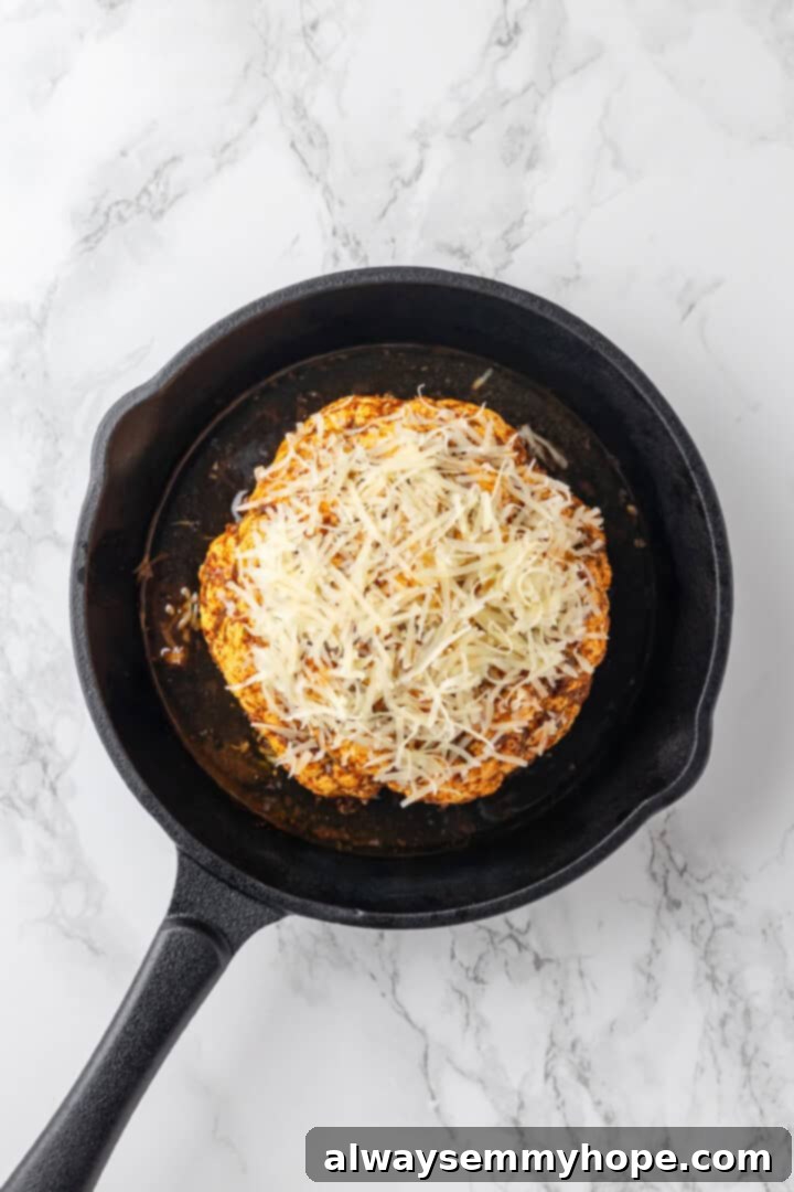 A whole roasted cauliflower in a cast iron skillet, now generously sprinkled with grated vegan Parmesan cheese, ready for its final bake.