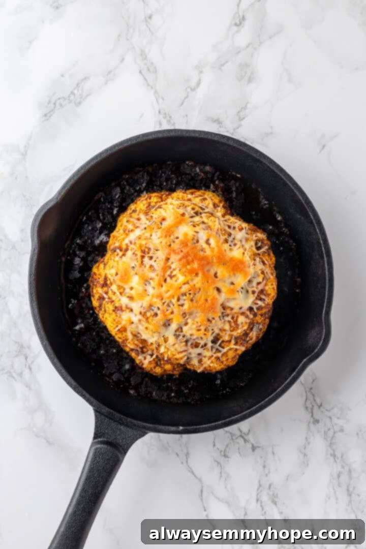 A whole roasted cauliflower, now perfectly golden brown with melted vegan cheese, resting in the skillet after being removed from the oven.