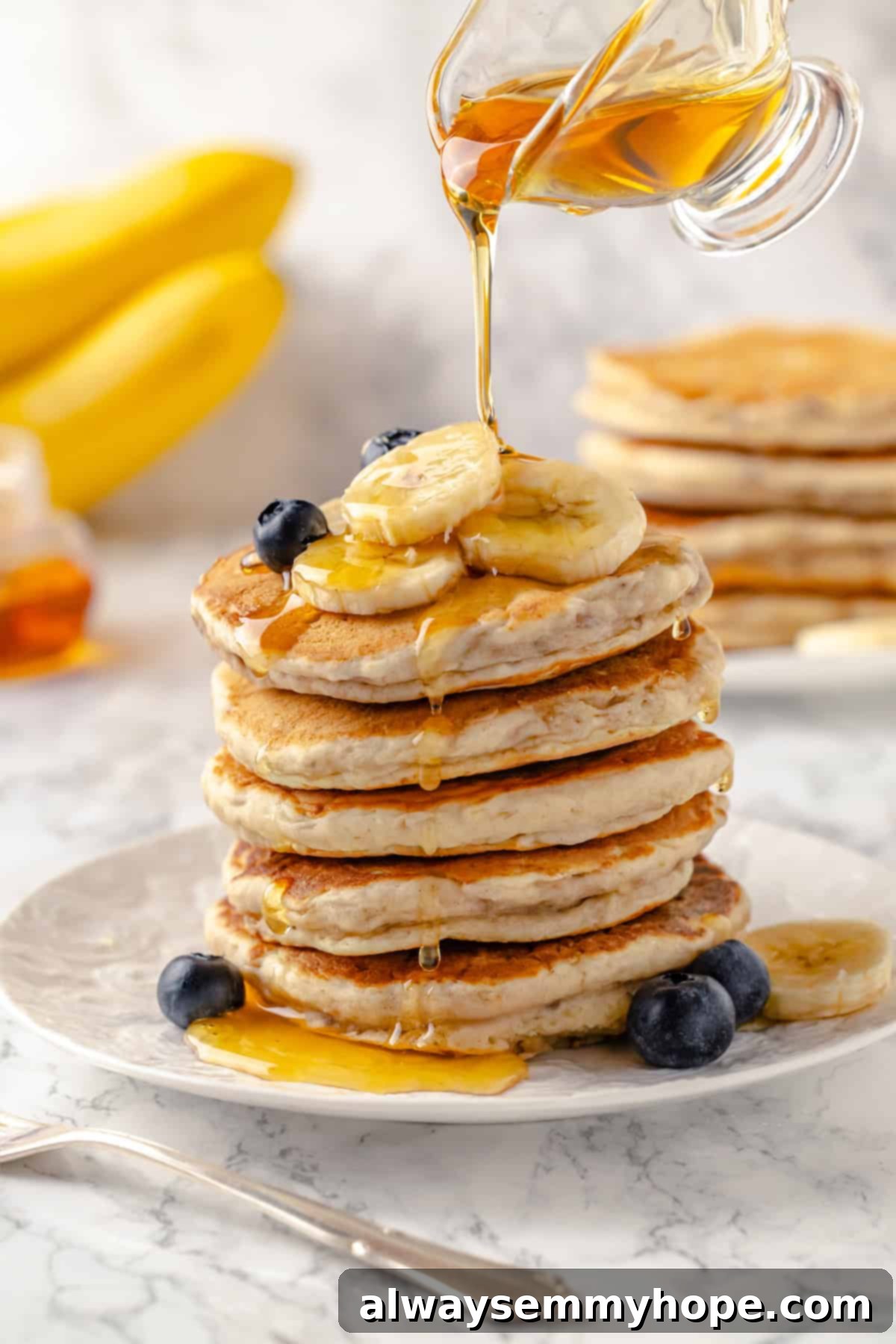 Pouring golden maple syrup onto a tall stack of fluffy banana pancakes, garnished with fresh berries