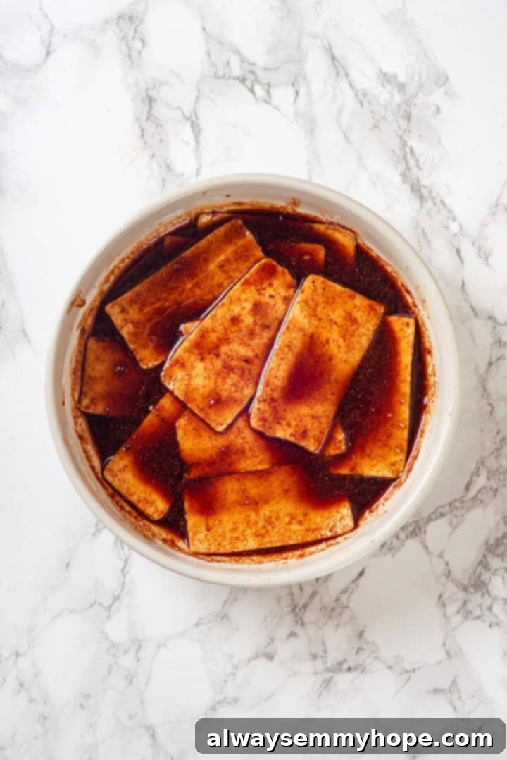 Marinating Tofu for Enhanced Flavor Overhead shot of thin tofu slices submerged and soaking in a savory marinade in a shallow dish, ensuring every piece is coated.
