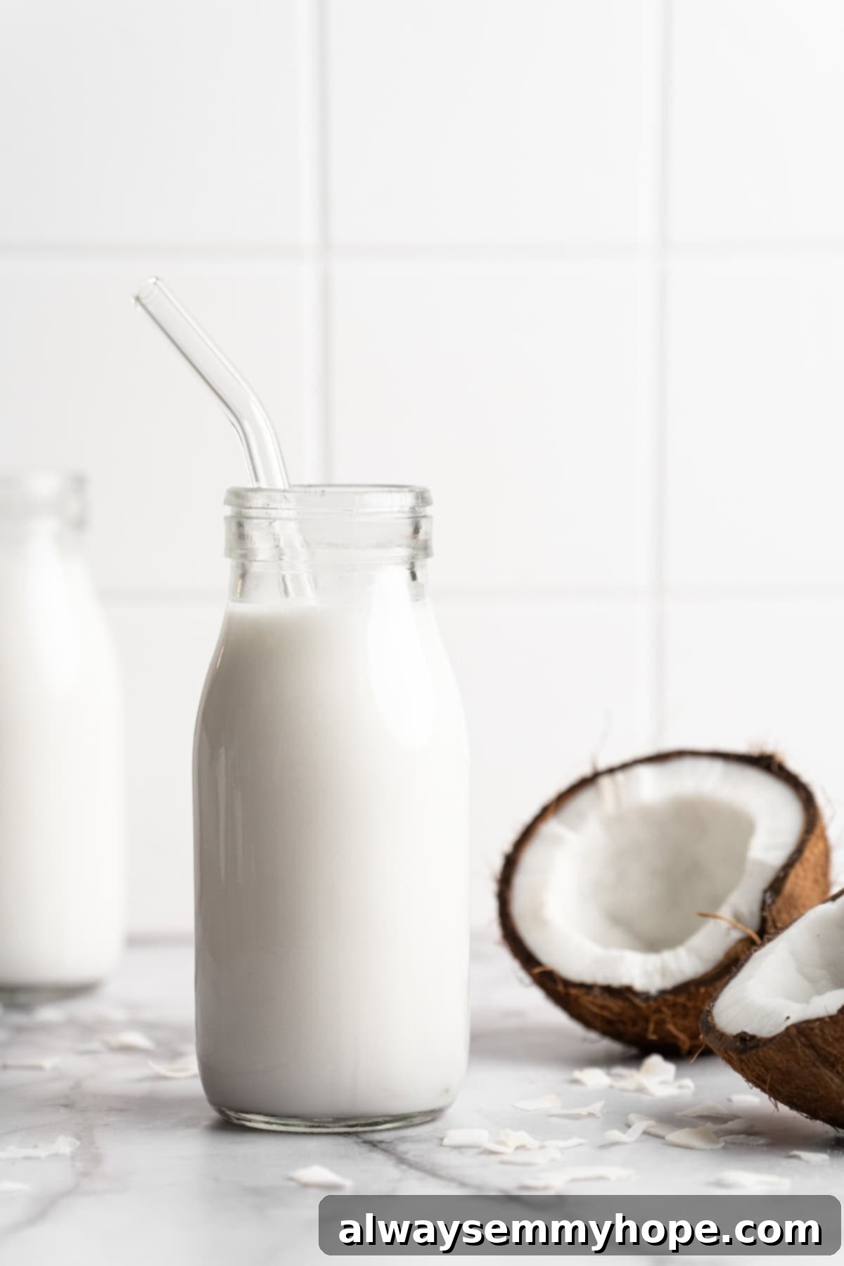 A glass bottle of homemade coconut milk with a reusable glass straw, placed on a light surface.