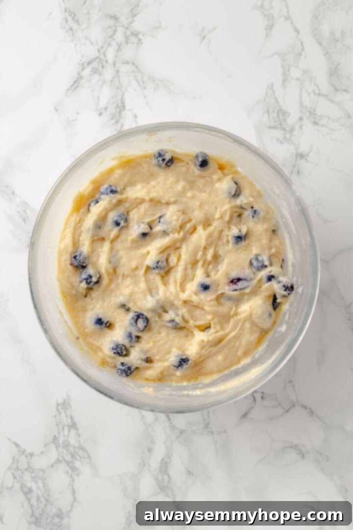 Gently fold blueberries into the batter. Overhead shot of the completed lemon blueberry Bundt cake batter in a large mixing bowl, showing the blueberries evenly folded in.