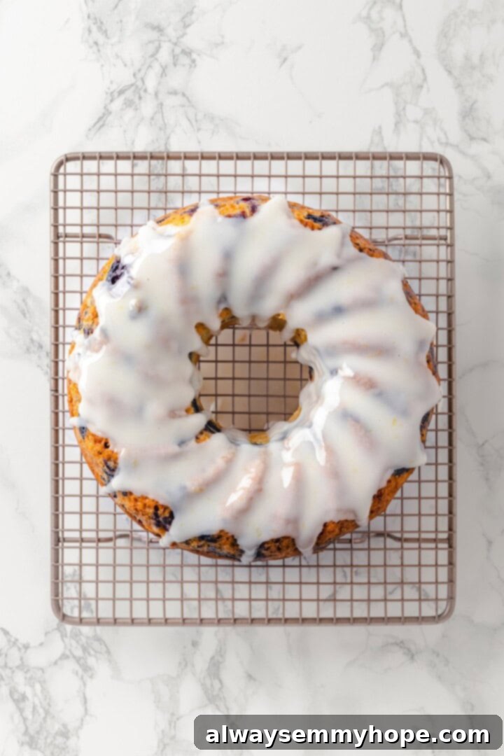 Drizzle the lemon glaze over the cooled cake. Overhead view of the cooled lemon blueberry Bundt cake on a wire rack, being drizzled with a thick, white lemon glaze.
