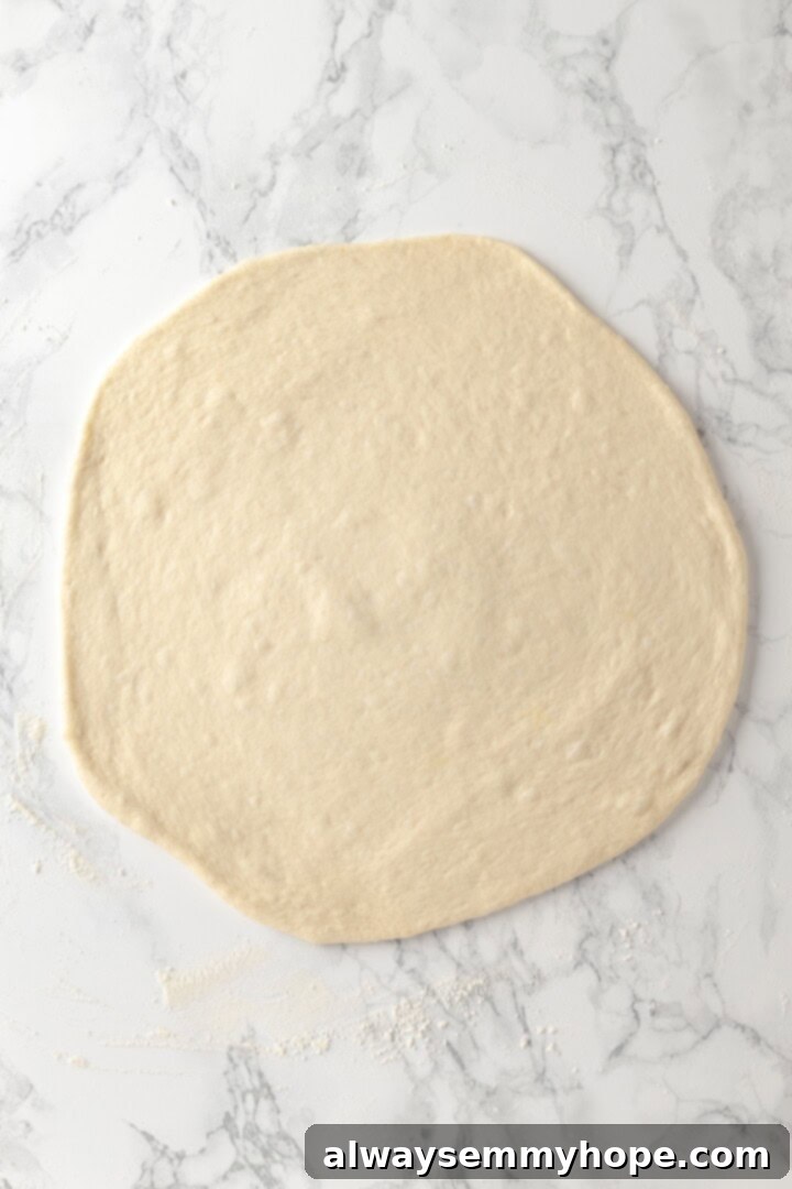 A creamy sauce makes this Alfredo sauce pizza taste indulgent, but it’s vegan! Plant-based chicken and bacon take this recipe over the top. Overhead view of pizza dough rolled out onto marble countertop, ready for topping.