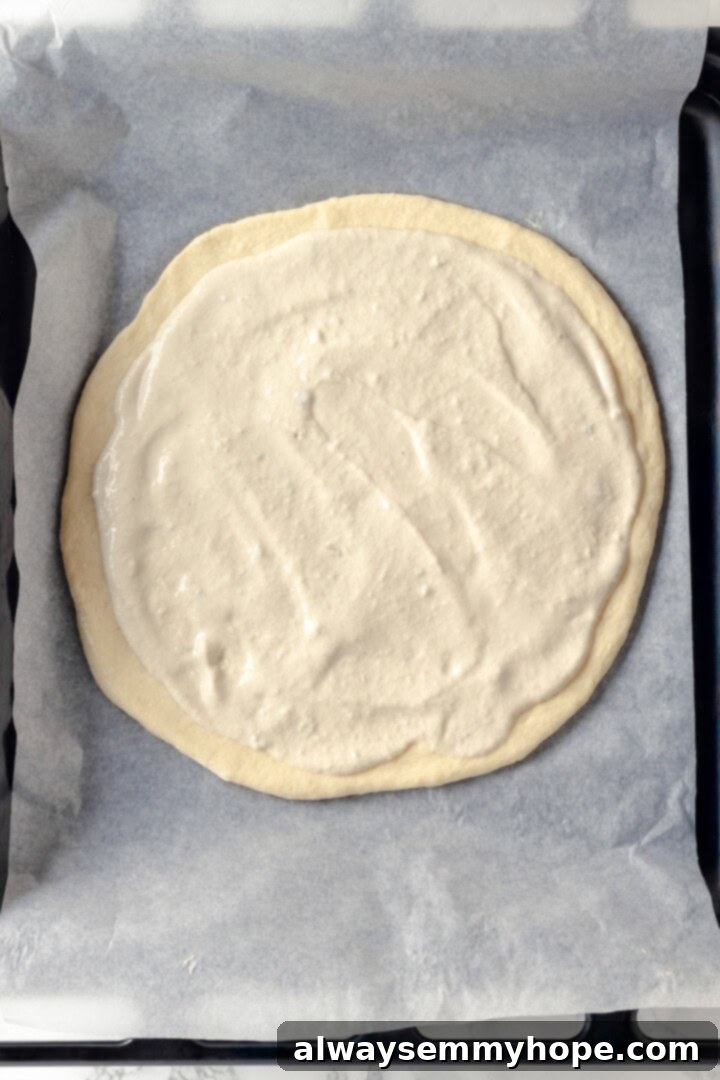 A creamy sauce makes this Alfredo sauce pizza taste indulgent, but it’s vegan! Plant-based chicken and bacon take this recipe over the top. Overhead view of an unbaked Alfredo sauce pizza on a pan, with the creamy sauce spread evenly.