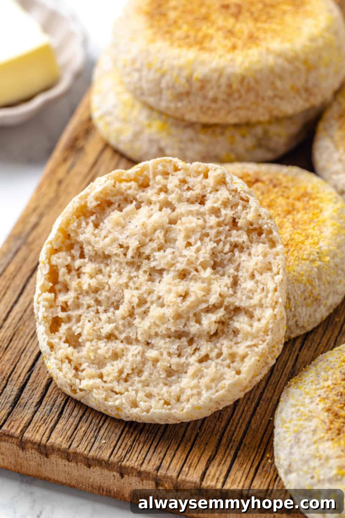 This homemade gluten-free English muffin recipe is tender, golden, and perfect for adding your favourite jam or spread. They're vegan too! Close-up of a sliced gluten-free English muffin half on a cutting board, revealing its airy interior and nooks and crannies, with more muffins in the background.