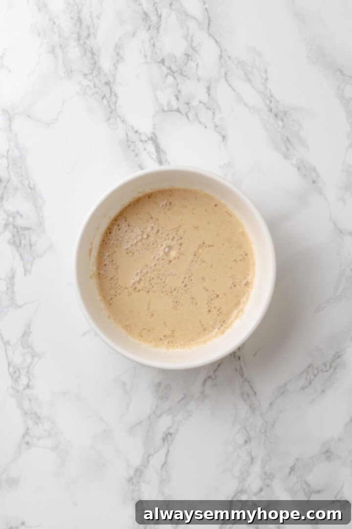 This homemade gluten-free English muffin recipe is tender, golden, and perfect for adding your favourite jam or spread. They're vegan too! Small bowl containing warm water, agave syrup, and instant yeast, showing foamy activation.