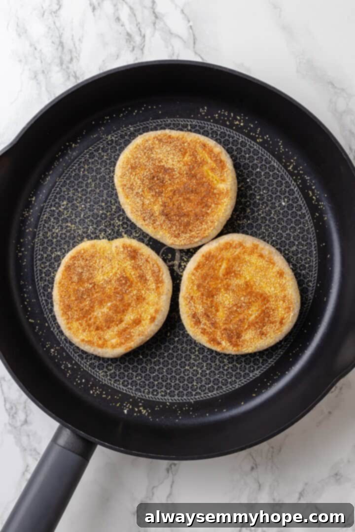 This homemade gluten-free English muffin recipe is tender, golden, and perfect for adding your favourite jam or spread. They're vegan too! Several gluten-free English muffins cooking in a hot cast-iron skillet, turning golden brown.