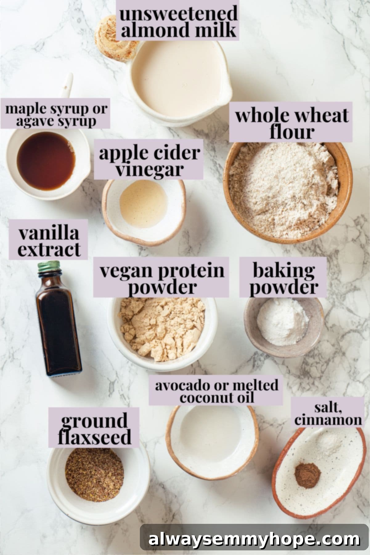 Overhead view of all ingredients for vegan protein waffles laid out on a kitchen counter, including almond milk, apple cider vinegar, whole wheat flour, protein powder, flaxseed, baking powder, cinnamon, salt, maple syrup, vanilla extract, and oil