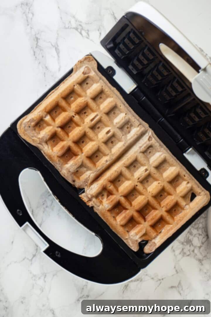 Overhead view of two golden-brown protein waffles cooking in an open waffle maker, showing their perfectly crisp edges and fluffy interior