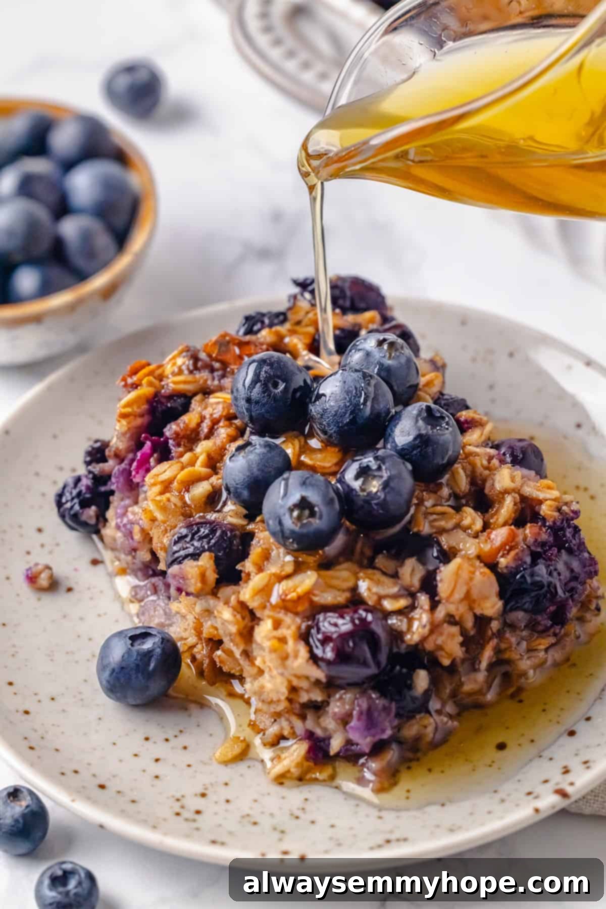 This baked oatmeal recipe is an easy meal prep breakfast! It's hearty and wholesome, with juicy berries and crunchy walnuts in every bite. Pouring maple syrup onto a portion of blueberry baked oatmeal on a white plate