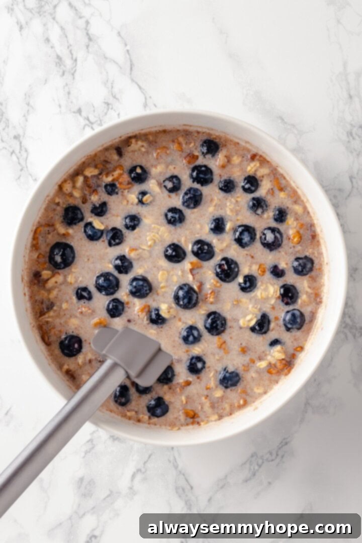 This baked oatmeal recipe is an easy meal prep breakfast! It's hearty and wholesome, with juicy berries and crunchy walnuts in every bite. Overhead view of oatmeal and blueberry mixture in a bowl with a rubber spatula, ready for baking