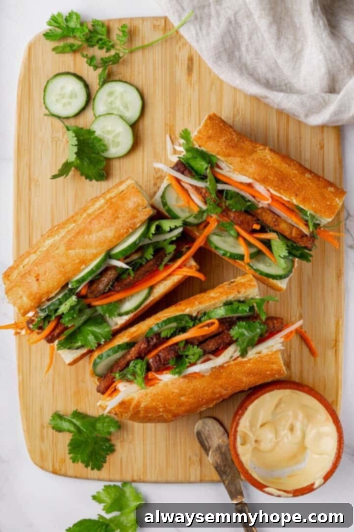Crispy air fried tofu is layered with crunchy pickled veggies on crusty baguette to make this fresh and flavourful vegan Tofu Banh Mi recipe! Three beautifully assembled vegan banh mi sandwiches with crispy tofu, pickled vegetables, and fresh herbs on a wooden cutting board