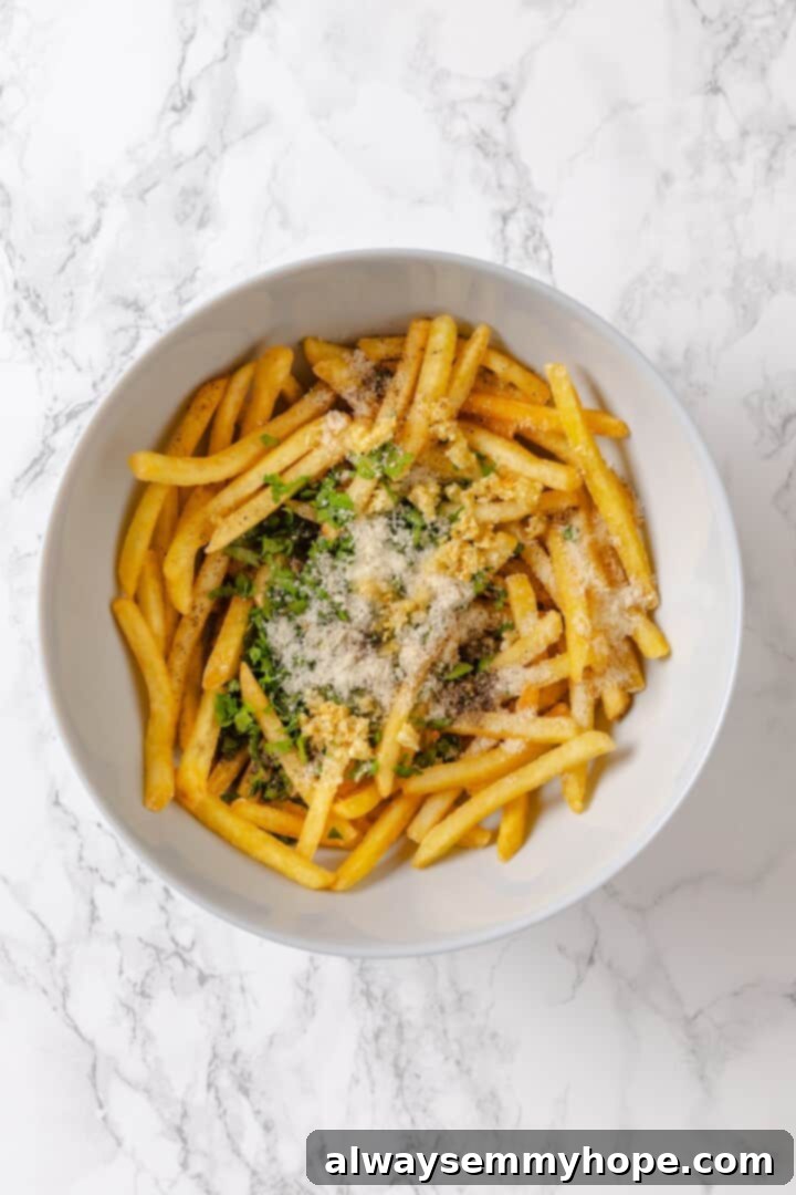 Whether you bake them or deep fry them, these crispy Garlic Fries are guaranteed to be a hit! They're cheesy, easy and packed with flavour. Overhead view of Parmesan, garlic, and parsley added to bowl of fries