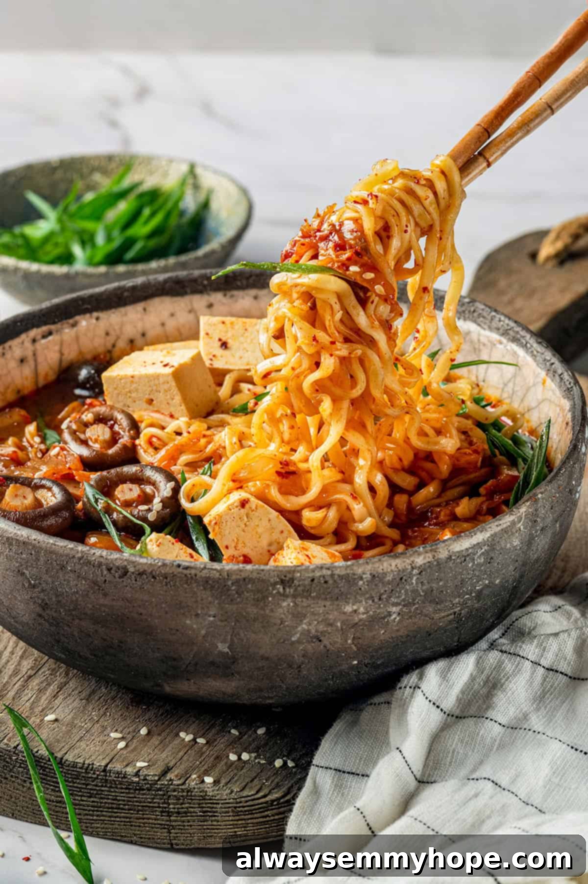 This spicy Kimchi Ramen is ridiculously easy, and the combination of tofu, kimchi, and shiitakes makes it a satisfying, flavour-packed meal! Chopsticks lifting a generous portion of noodles and kimchi from a bowl of spicy vegan kimchi ramen, showcasing the rich texture and ingredients.