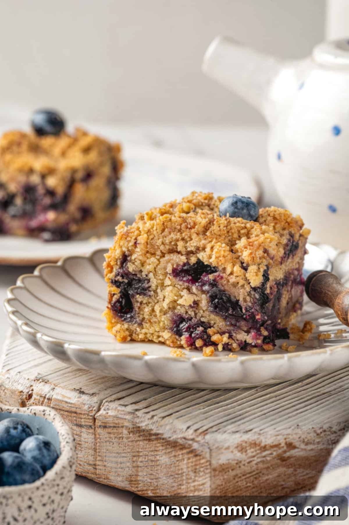 A little bit like coffee cake, this Blueberry Buckle is loaded with berries, then topped with brown sugar cinnamon streusel. So delicious! A slice of vegan blueberry buckle on a white plate with a blurry teapot in the background, ready for serving.