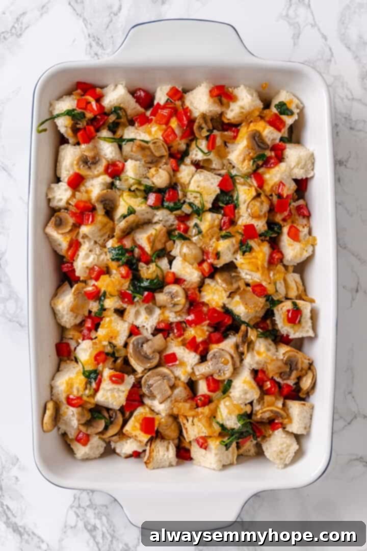 A baking dish filled with layers of cubed bread and sautéed vegetables, prepared for the next step of the strata recipe.