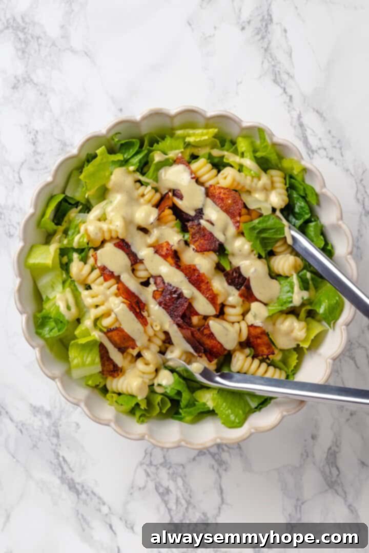 This vegan Caesar Pasta Salad is savoury, tangy and garlicky, with crunchy baked chickpeas, crisp romaine and tender pasta. Delicious! Tongs tossing caesar pasta salad in mixing bowl.
