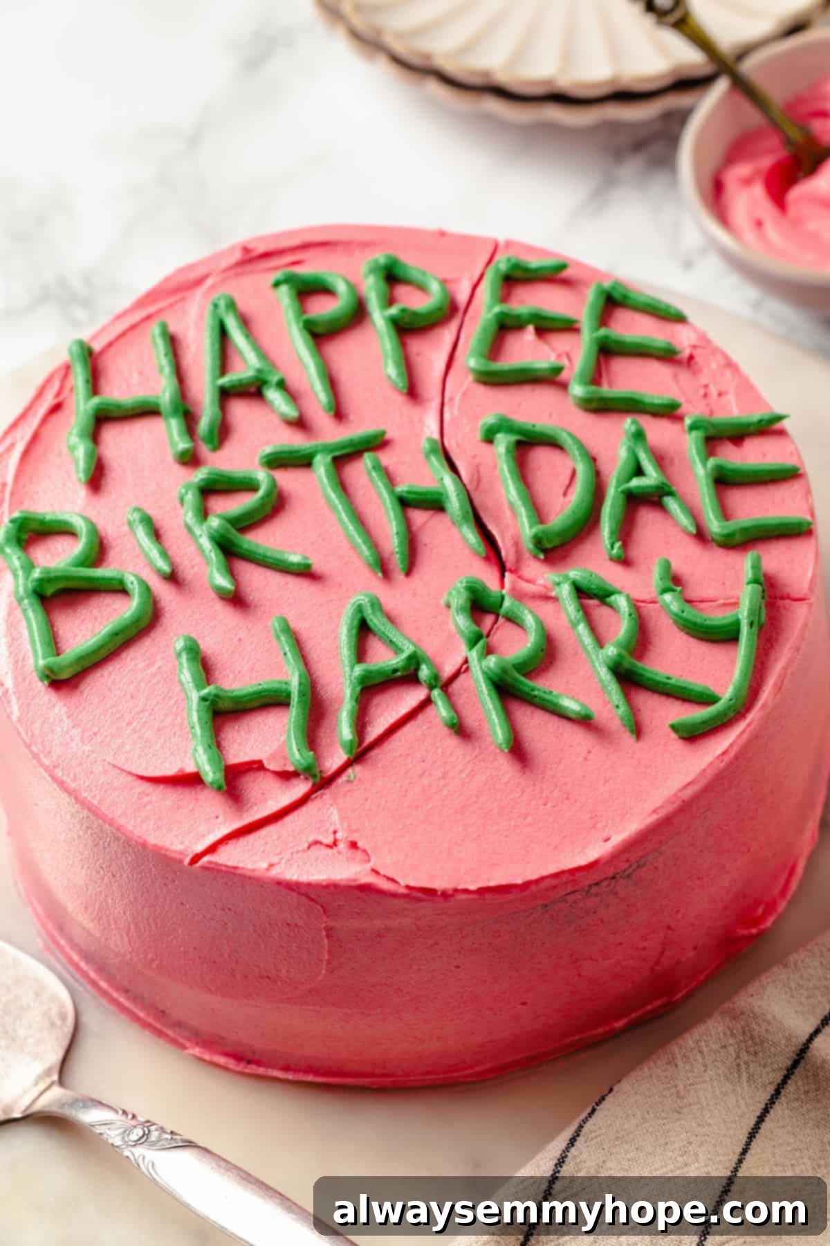 Homemade Harry Potter cake that reads Happee Birthdae Harry. The cake is round with pink frosting and green letters spelling 'Happee Birthdae Harry.' It sits on a light wooden surface.