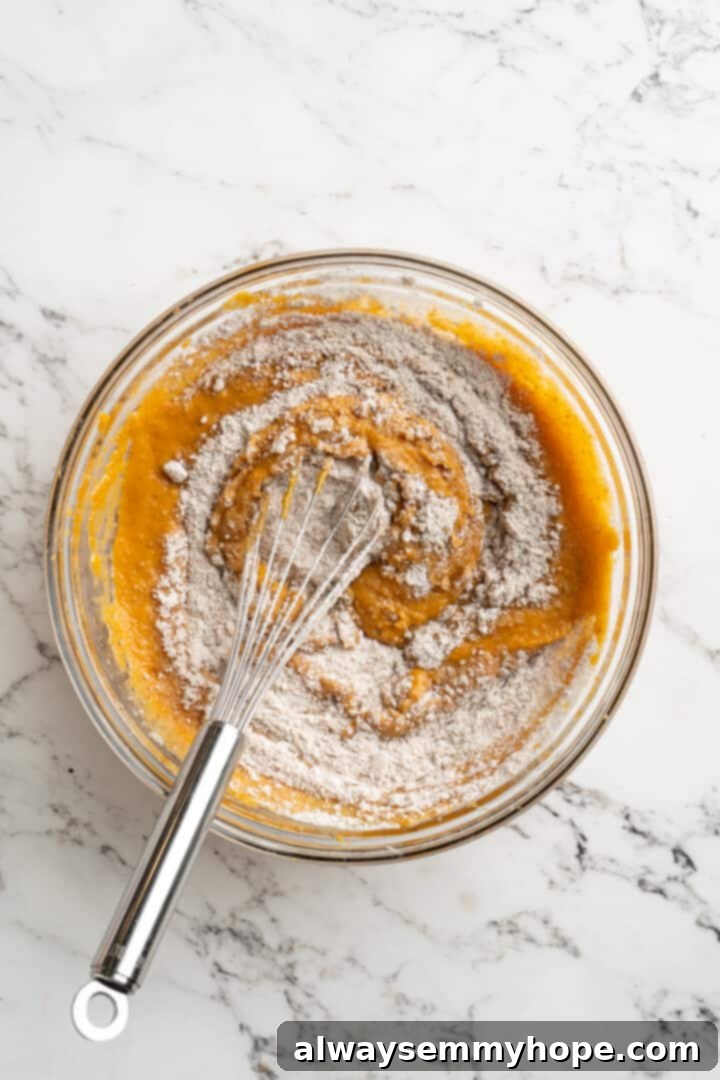 Overhead view of a whisk blending flour into a bowl of pumpkin puree and wet ingredients, creating a thick batter.