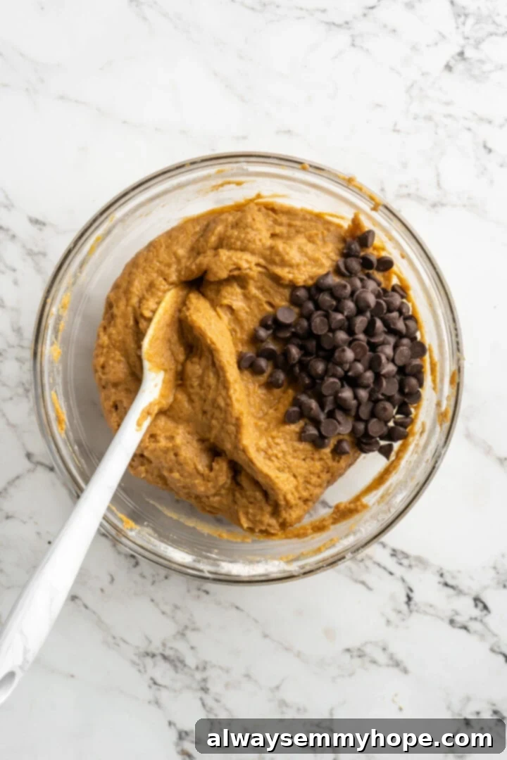 Overhead view of a pile of vegan chocolate chips resting on top of pumpkin muffin batter in a bowl, with a spatula ready to fold them in.