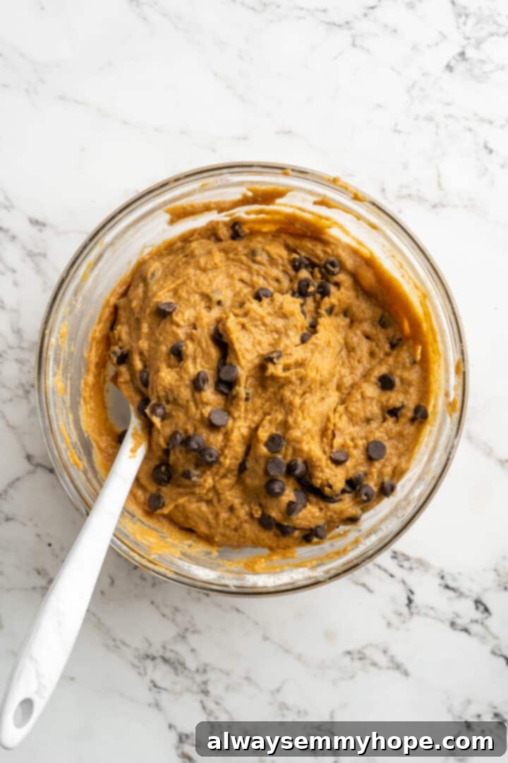 Overhead view of a mixing bowl with pumpkin chocolate chip muffin batter and a spatula, showing the chocolate chips evenly mixed throughout.