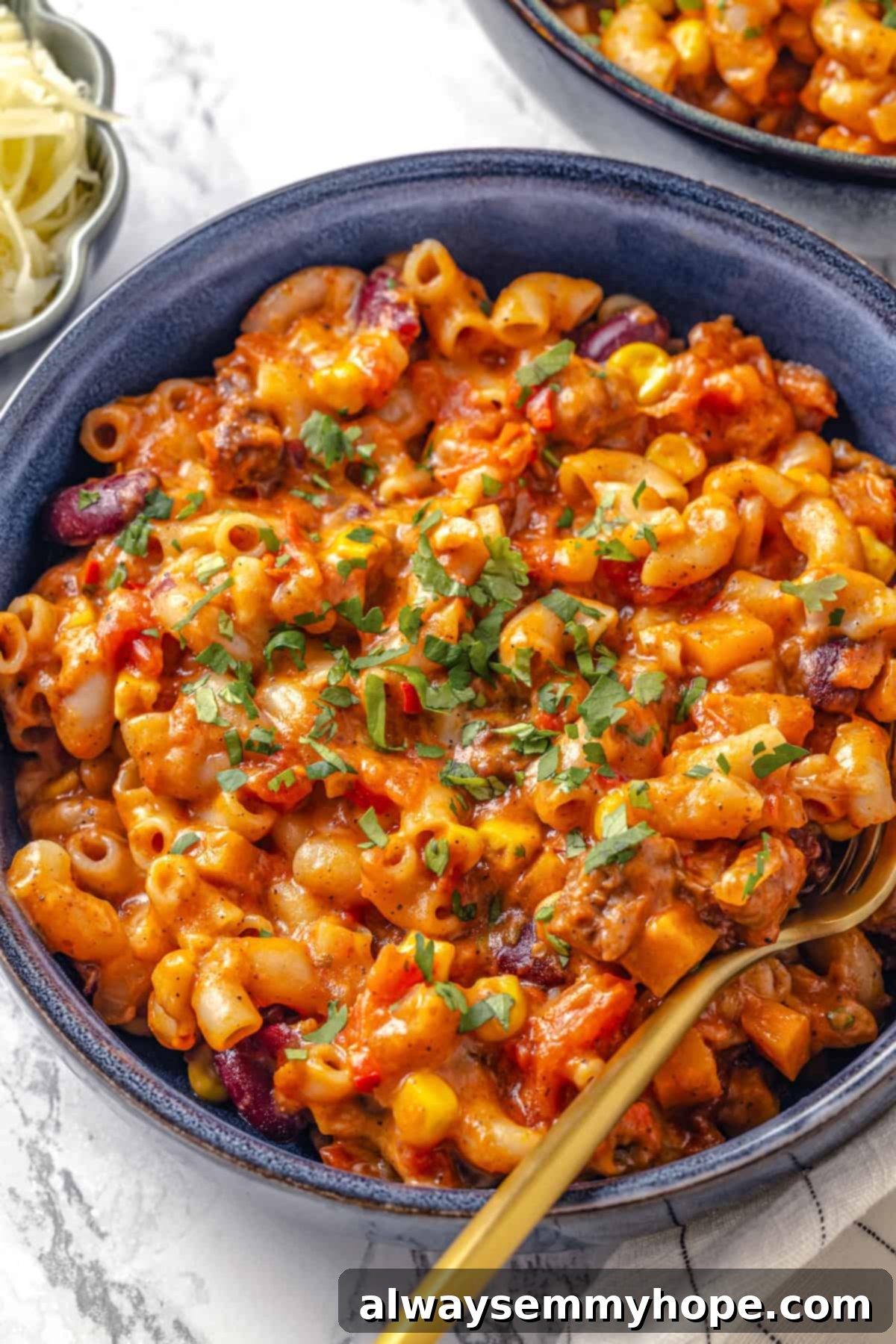 This Chili Mac and Cheese recipe is loaded with vegan ground beef, beans and lots of plant-based cheese. A kid-friendly comfort food dinner! A close-up shot of a bowl of steaming vegan chili mac and cheese, garnished with fresh cilantro, with a fork ready to dig in. Rich, vibrant colors highlight the chili, pasta, and melty cheese.