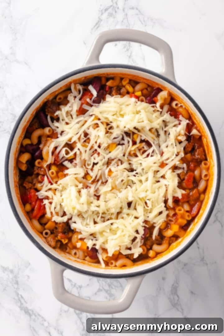 This Chili Mac and Cheese recipe is loaded with vegan ground beef, beans and lots of plant-based cheese. A kid-friendly comfort food dinner! Shredded vegan cheese being added to the hot chili mac, ready to melt into a creamy sauce.